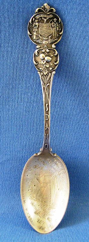 Souvenir Mining Spoon Hecla Mine.JPG - SOUVENIR MINING SPOON HECLA MINE BURKE IDAHO - Sterling silver souvenir spoon, HECLA MINE BURKE engraved in bowl above engraved depiction of mine structure, handle finial has a coat of arms of the State of Idaho in a floral design on both sides and IDAHO written on the neck of the stem, IH on a triangular flag manufacturer’s mark and STERLING in the mold on the back, 5 in. long, 15.1 grams  (Burke was once a thriving silver, lead and zinc mining community located about 7 miles northeast of Wallace, ID.  It’s now a ghost town at the upper end of a string of seven historic mining camps that ran between Gem and Burke in Idaho’s Shoshone County.  Today the town still shows the remains of the massive brick buildings that were once the Hecla Mining Company offices and mine, whose history is intertwined with the town.  With the discovery of rich silver ore, Burke was founded in 1884 as mines and mills were built on the surrounding hillsides of the town’s narrow canyon.  Wooden buildings and a railroad followed within three years and the boomtown was on.   The Hecla Mine was originally discovered by James Toner on May 5, 1885.  His 20-acre silver-lead claim was one of many in the rich Coeur d’Alene Mining District.  He sold it and the mine changed hands several times before it was purchased for $150 by a group of investors in 1891.  These investors led by Amasa B. Campbell, Patsy Clark and John Finch, founded the Hecla Mining Company and incorporated it in the state of Idaho on Oct. 14, 1891.  After seven years of leasing the property, they recapitalized, booted the leasers out and began mining themselves.  They reorganized the company in Washington state on July 12, 1898, electing Amasa B. Campbell as its president and capitalizing the company to $250,000.  In 1900 they were well on their way to profitability, having built up a large surface plant to process their ore.  By the end of 1900, the Hecla Mine produced $229,500 worth of ore.  Just over 15 years after its discovery, the mine became one of the top producers in Burke.  In Oct. 1904, Hecla’s offices were moved from Spokane to Wallace, ID where they remained for over 80 years. Through the economic ups and downs of the first two decades of the 20th Century, the Hecla Mine was a true bright spot, pumping out its silver, lead and zinc treasures.  In 1922, the neighboring Star Mine was purchased by the Hecla Mining Company, and the two mines were then connected by a two-mile long underground tunnel.  Unfortunately, the town of Burke was a disaster waiting to happen.  It was a compact cluster of wooden buildings jammed into the bottom of a narrow canyon.  Because of the narrowness of that hundred-yard wide canyon, the railroad ran through the middle of town.  Most towns had main streets.  Burke had a railroad.  Because of this narrowness, most of the town’s buildings crowded up against the railroad, or spanned over it.  Very few wooden mining towns with Burke’s character escaped the ravages of fire.  On July 13, 1923, the inevitable happened.  Fire ripped through the wooden mass of buildings, quickly burning nearly three-quarters of the town, and reducing the Hecla Mine office and milling complex to ashes.  By the early part of 1925, the town was rebuilt of brick and it was business as usual.  The good times were short lived and the Great Depression and the collapse of zinc prices in the 30s led to the closure of the Star Mine.  The Hecla Mine held on to till 1944 when it too closed after producing nine million tons of ore, yielding 41 million ounces of silver, 732,000 tons of lead and 41,275 tons of zinc, worth some $81 million.  Hecla reopened the Star Mine and continued to expand its operations by purchasing additional mining properties.  Today Hecla Mining Company is the largest primary silver producer in the U.S. through its primary mines, the Greens Creek Mine in Alaska and the Lucky Friday Mine in Idaho.  The company’s expected 2014 silver production is between 9.5 and 10 million ounces.) 